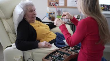 Yaşlı bir kadın, sağlık hizmetlerinde çalışan bir bakıcı ile keyifli bir mücevher oturumunda rahat bir koltukta dinlenir..