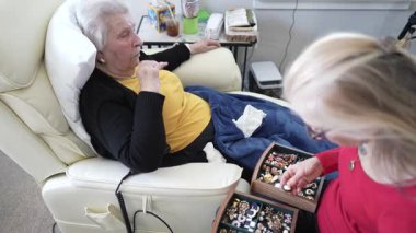 Yaşlı bir kadın, bakıcı olarak bir sandalyede rahatlar ve ev sağlığı merkezindeki bir koleksiyondan mücevher seçmesine yardım eder..