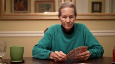 Yaşlı bir kadın evindeki masasında kart oynamaktan hoşlanır. Kartlarını tutarken odaklanmış ve meşgul görünüyor..