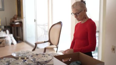 Olgun bir kadın, kutulardan antika porselenleri dikkatlice açar. Aile yemeklerini ve geçmişteki anılarını hatırlar..