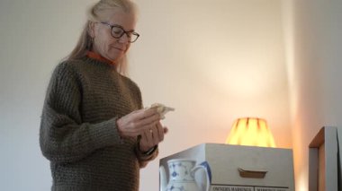 Olgun bir kadın aile yadigarlarını bulmak için kutuları açar ve güneşli bir günde oturma odasında porselen parçalarını inceler..