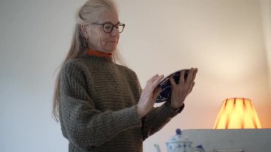 Olgun kadın aile yadigârlarını inceliyor ve evdeki parlak yaşam alanındaki kutuları açarken anılarını paylaşıyor.