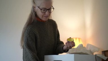 Olgun bir kadın oturma odasında aile yadigârlarıyla dolu kutuları açar ve her parçaya özenle ve neşeyle bakar..