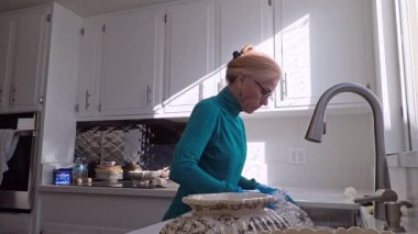 Olgun bir kadın, parlak mutfağında antika porselen kaseleri ve tabakları dikkatlice yıkıyor. O çalışırken güneş ışığı odayı dolduruyor..