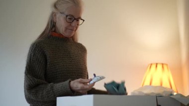 Mature woman opens boxes to reveal family heirlooms and antiques while exploring her inheritance at home in the living room.
