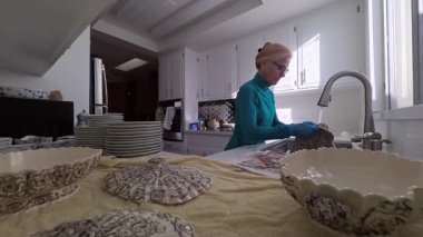 Olgun bir kadın, aile geleneklerini sürdürürken antika porselen kaselerini ve tabaklarını parlak bir mutfakta dikkatlice yıkıyor..