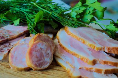 İnce dilimlenmiş et ve ahşap bir kesme tahtasının üzerinde taze dereotu ve maydanozla süslenmiş ev yapımı sosis çeşitleri. İştah açıcı pembe dilimler, altın kabuk ve parlak yeşil otlar Polonya atmosfer geleneğini vurguluyor