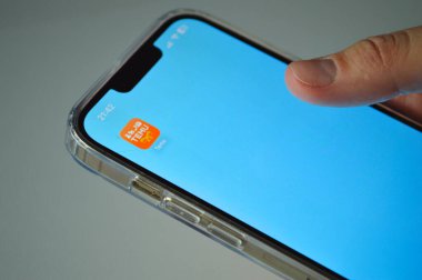 Açık mavi bir ev ekranında Temu uygulama simgesini gösteren bir akıllı telefonun yakın görüntüsü, şeffaf bir koruyucu çantadaki aygıt ve en üstte gösterilen saat.