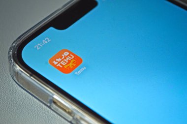 Açık mavi bir ev ekranında Temu uygulama simgesini gösteren bir akıllı telefonun yakın görüntüsü, şeffaf bir koruyucu çantadaki aygıt ve en üstte gösterilen saat.