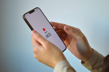 Tarafsız bir arkaplanda IKO mobil bankacılık uygulamasının logosunu gösteren akıllı telefonu tutan ellerinizi kapatın. Maliye, dijital ödemeler ve Polonya bankacılık sektörü ticari kullanımı için ideal