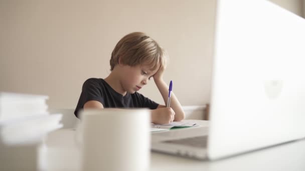 Uma criança faz trabalhos de casa usando um laptop enquanto está sentada em casa na cozinha à mesa. Tecnologia Online. — Vídeo de Stock