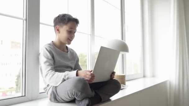 Jonge jongen werkt op een laptop computer terwijl ontspannen op de vensterbank thuis in een helder appartement. — Stockvideo