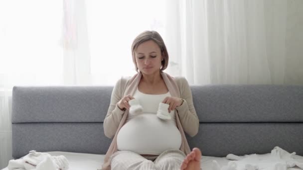 Mladá těhotná žena s malými botičkami sedící doma na pohovce. Očekávající matka se připravuje na porod. — Stock video