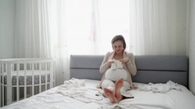 Mutlu hamile kadın evdeki kanepede otururken bebek kıyafetlerine şefkatle bakıyor. Müstakbel anne, doğmadan önce doğmamış bebek için alışveriş yapıyor.