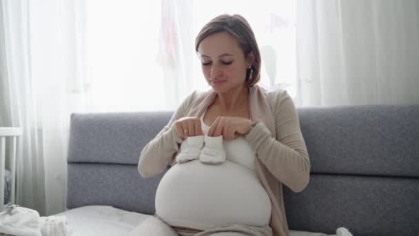 Young pregnant woman with small baby shoes sitting on sofa at home. Expectant mother is preparing for childbirth. — Stock Video
