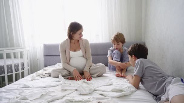 Gravid kvinna sitter på sängen med två söner och förbereder saker för födelsen av ett barn. — Stockvideo