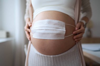 Hamile anne karnına yüz maskesi takarak fetüs ya da bebeği koronavirüsten, covid19, kirlilik, grip ya da salgından korur. Hamile kadın evde karantina altında.