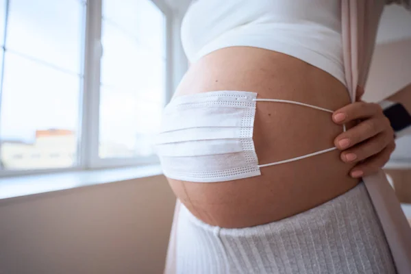 Hamile anne karnına yüz maskesi takarak fetüs ya da bebeği koronavirüsten, covid19, kirlilik, grip ya da salgından korur. Hamile kadın evde karantina altında.
