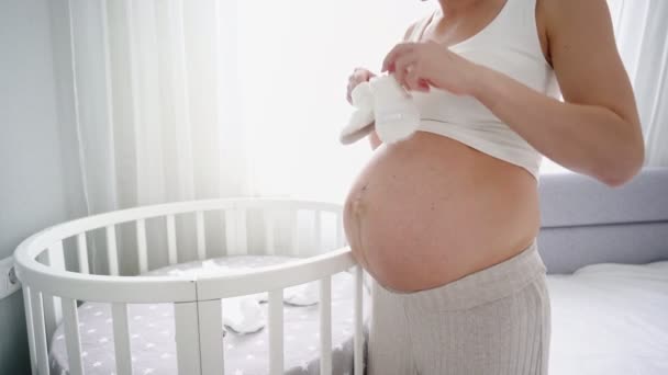 Mujer embarazada joven con zapatos de bebé pequeños sentados en el sofá en casa. La madre embarazada se está preparando para el parto. — Vídeo de stock