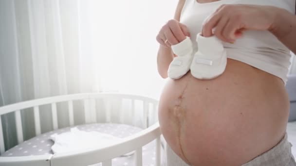 Junge schwangere Frau mit kleinen Babyschuhen sitzt zu Hause auf dem Sofa. Werdende Mutter bereitet sich auf Geburt vor. — Stockvideo