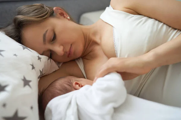 Güzel bir kadın bebeğini yatağın üzerinde rahat bir evde emziriyor..