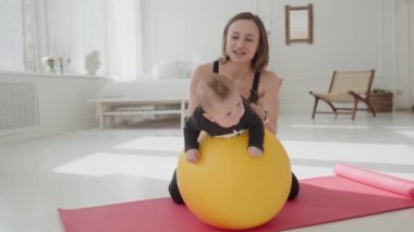 Annesi bacaklarını tutarken, spor yapan tatlı bir çocuk spor yapıyor. Evde eğitim, sağlıklı çocukluk ve spor geliştirme süresince, zayıf formda bir kadın, yeni yürümeye başlayan bir çocuğu top üstünde sallıyor..