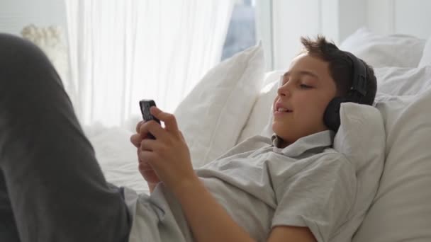11歳の男の子がベッドに横になり、ヘッドフォンで音楽を聴く — ストック動画