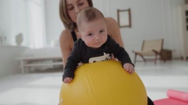 Annesi bacaklarını tutarken, spor yapan tatlı bir çocuk spor yapıyor. Evde eğitim, sağlıklı çocukluk ve spor geliştirme süresince, zayıf formda bir kadın, yeni yürümeye başlayan bir çocuğu top üstünde sallıyor..