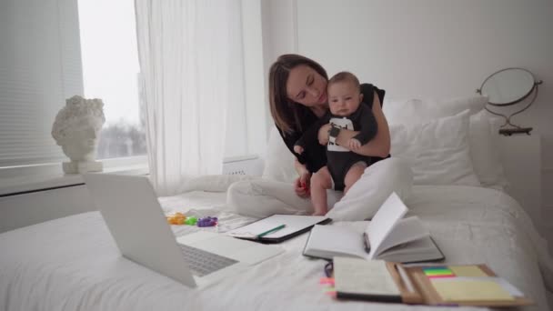 Joven madre freelancer con bebé hijo sentado en la cama y tratando de trabajar en el ordenador portátil — Vídeo de stock