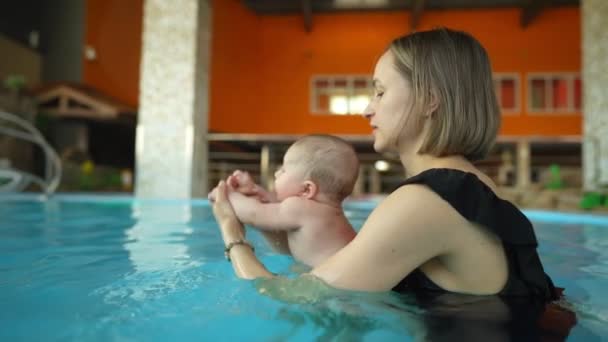 Mutter bringt ihrem Baby das Schwimmen in einem geschlossenen öffentlichen Schwimmbad bei. Das Kind schwingt auf einer künstlichen Welle im Aquapark — Stockvideo