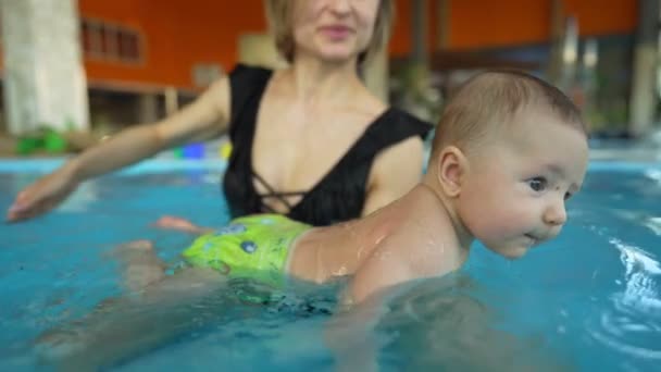 Madre enseña a su bebé a nadar en una piscina pública cerrada. El niño se balancea sobre una ola artificial en el parque acuático — Vídeo de stock