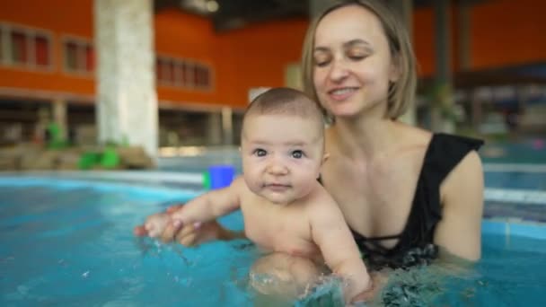 Madre enseña a su bebé a nadar en una piscina pública cerrada. El niño se balancea sobre una ola artificial en el parque acuático — Vídeos de Stock