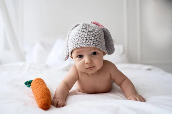 Bebê quatro meses de idade em um traje de coelho com uma cenoura de malha que coloca na cama branca — Fotografia de Stock