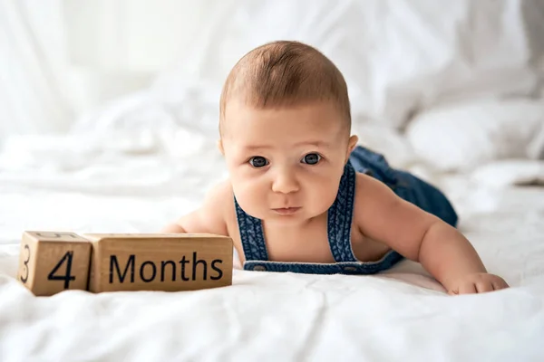 Adorable four month old baby boy in denim dungarees lying on white bed — Stock Photo, Image