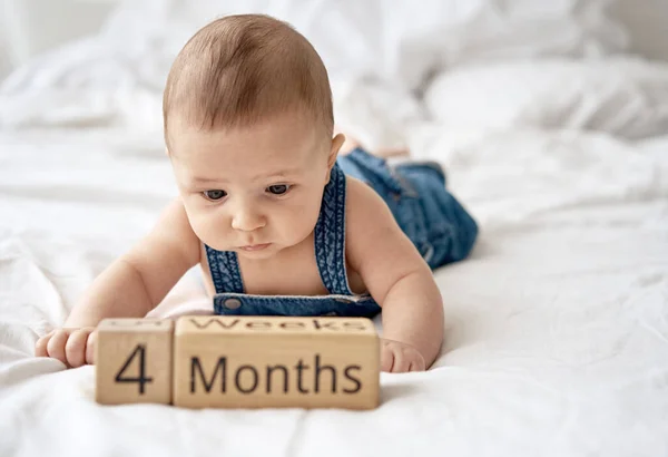 Adorable four month old baby boy in denim dungarees lying on white bed — Stock Photo, Image