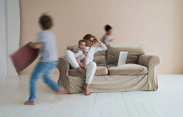 Joven madre soltera trabajando en el portátil en el loft sentado en el sofá mientras sus hijos corren a su alrededor y gritando. — Foto de Stock