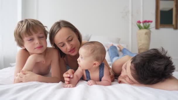 Mamma och tre söner har kul på en vit säng. Lyckligt familjebegrepp — Stockvideo