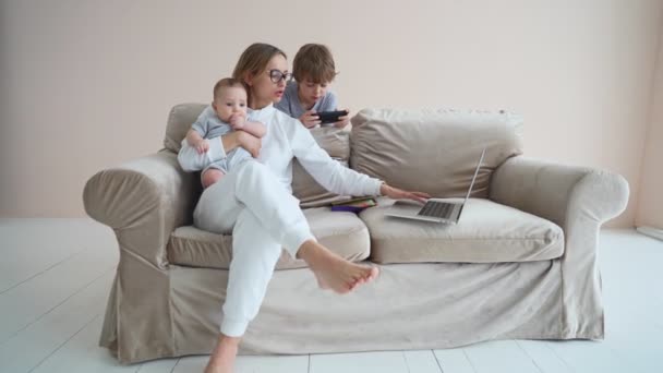 Trevlig ung mamma frilansare arbetar på datorn sitter på en soffa med söt baby spädbarn hemma. Vacker mamma studerar online, med hjälp av bärbar programvara, surfing information i nätbutik. — Stockvideo