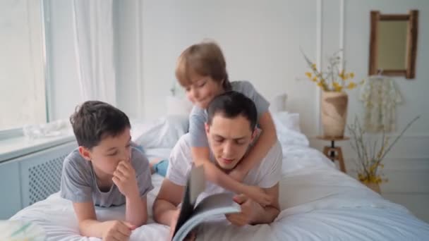 De vader brengt tijd door met de kinderen. Jonge vader bladert door een tijdschrift met kinderen liggend op het bed — Stockvideo