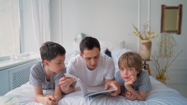 Отец проводит время с детьми. Молодой отец листает журнал с детьми, лежащими на кровати — стоковое видео
