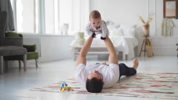 Ayah muda yang bahagia berbaring di atas karpet lantai, mengangkat bayi laki-laki yang menggemaskan udara, bermain pesawat atau membayangkan perjalanan bersama di ruang tamu, konsep hari ayah yang bahagia. — Stok Video