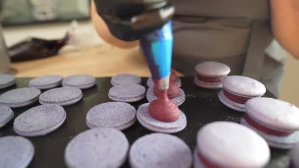 Prozess der Herstellung von Makronen zu Hause. Aufnahme von Händen einer Konditorin, die weißen Macaron mit Ganache in der Hand hält und rote Fruchtmarmelade aus der Tüte presst. Naturteigmakronen — Stockvideo