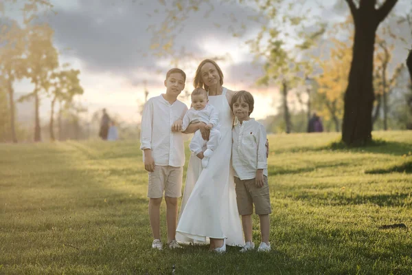 Mamma och tre barn kramas. Glad familj sitter utomhus: kvinna och två bror barn pojkar och söt liten småbarn pojke. — Stockfoto