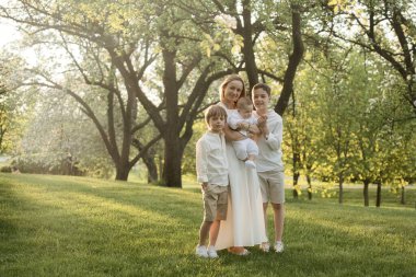 Anne ve üç çocuk sarılıyor. Mutlu bir aile dışarıda oturuyor: bir kadın ve iki kardeş çocuklar ve küçük şirin bir çocuk..