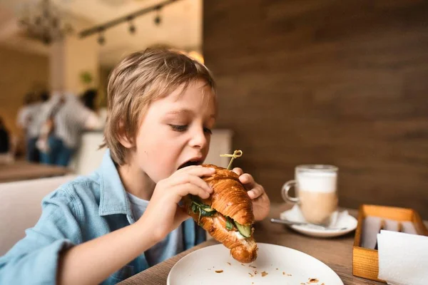 Feliz chico lindo comiendo croissant fresco, sentado a la mesa en la cafetería de la ciudad, con taza de papel de cacao — Foto de Stock
