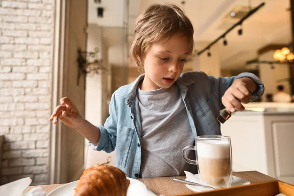 Feliz chico lindo comiendo croissant fresco, sentado a la mesa en la cafetería de la ciudad, con taza de papel de cacao — Foto de Stock