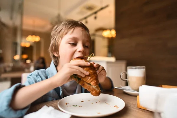 Happy cute boy eating fresh croissant, sitting at the table in city cafe, with paper cup of cocoa — Stock Photo, Image