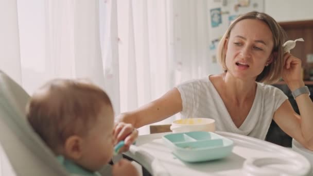 Baby zit aan een kindertafeltje. Mama voedt het kind met pap. Moeder geeft baby voedsel van een lepel. — Stockvideo