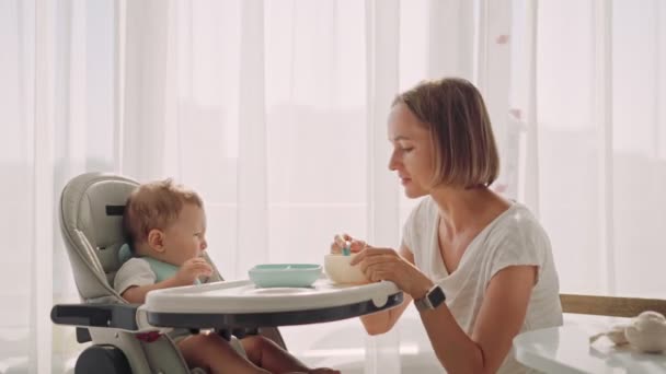 El bebé se sienta en una mesita de niños. Mamá alimenta al niño con gachas. Madre da comida de bebé de una cuchara. — Vídeo de stock