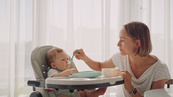 婴儿坐在孩子们的小桌旁。妈妈用粥喂孩子.妈妈用勺子喂婴儿. — 图库视频影像